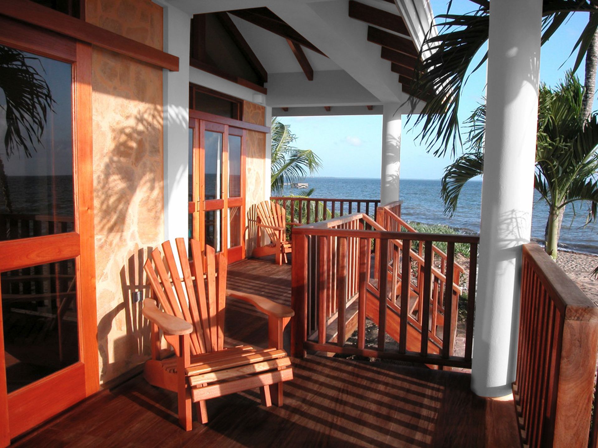 A wooden deck or balcony overlooking the ocean. A wooden Adirondack chair sits on the deck, and there are stairs leading down to the beach. Palm trees are visible in the background. The sky is blue and sunny.