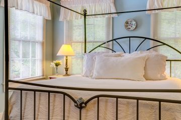 A cozy bedroom featuring a metal canopy bed, soft white bedding, and natural light from large windows.