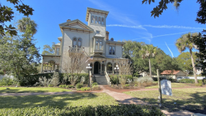A historic mansion surrounded by greenery and palm trees under a clear blue sky.