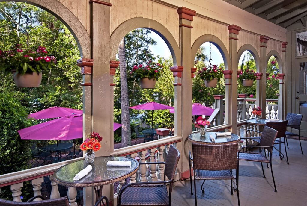 A charming outdoor dining area featuring glass tables, wicker chairs, and vibrant potted flowers under archways, with pink umbrellas in the background.