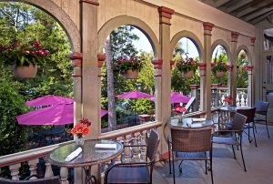 A charming outdoor dining area with arched columns, tables, and purple umbrellas surrounded by greenery.