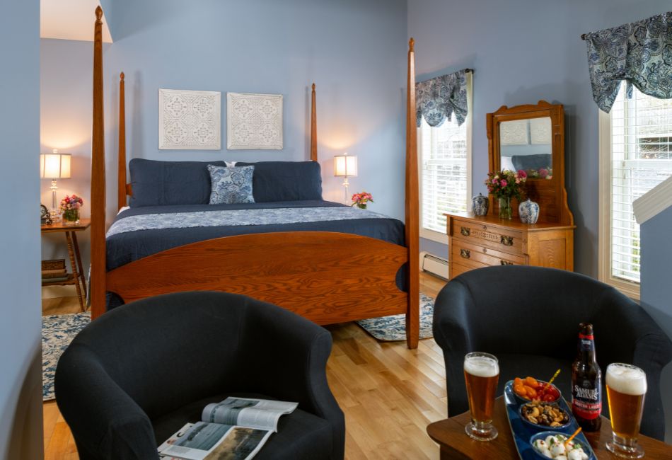 A cozy bedroom featuring a four-poster bed, two armchairs, a decorative mirror, and a side table with food and drinks.