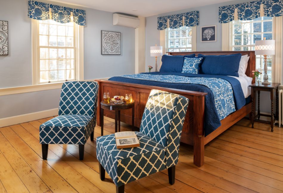 A cozy bedroom featuring a wooden bed with blue bedding, two patterned chairs, and natural light from large windows.
