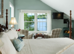 A cozy bedroom with a view of a river, featuring a large bed, two armchairs, and a small table set with snacks and drinks.