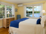 A cozy, sunlit bedroom with a blue and white bedspread, dresser, and views of a lake through large windows.