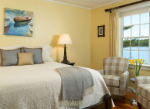 A cozy bedroom with a bed, accent pillows, a lamp, and a window overlooking a body of water, adorned with a painting of boats.