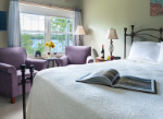 A cozy bedroom featuring a bed with a quilt, an open book, two chairs, a window with a view, and decorative elements.
