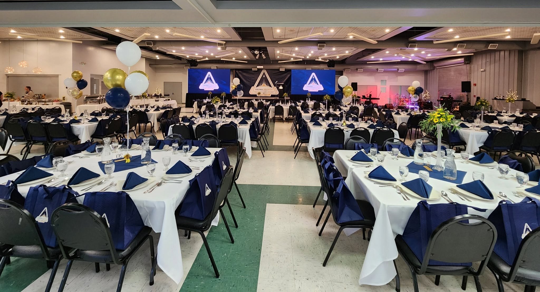 The Chincoteague Center’s main hall set with round tables in white linens and navy napkins, gold and navy balloons, and a stage with screens showing the Virginia Spaceport Authority logo.