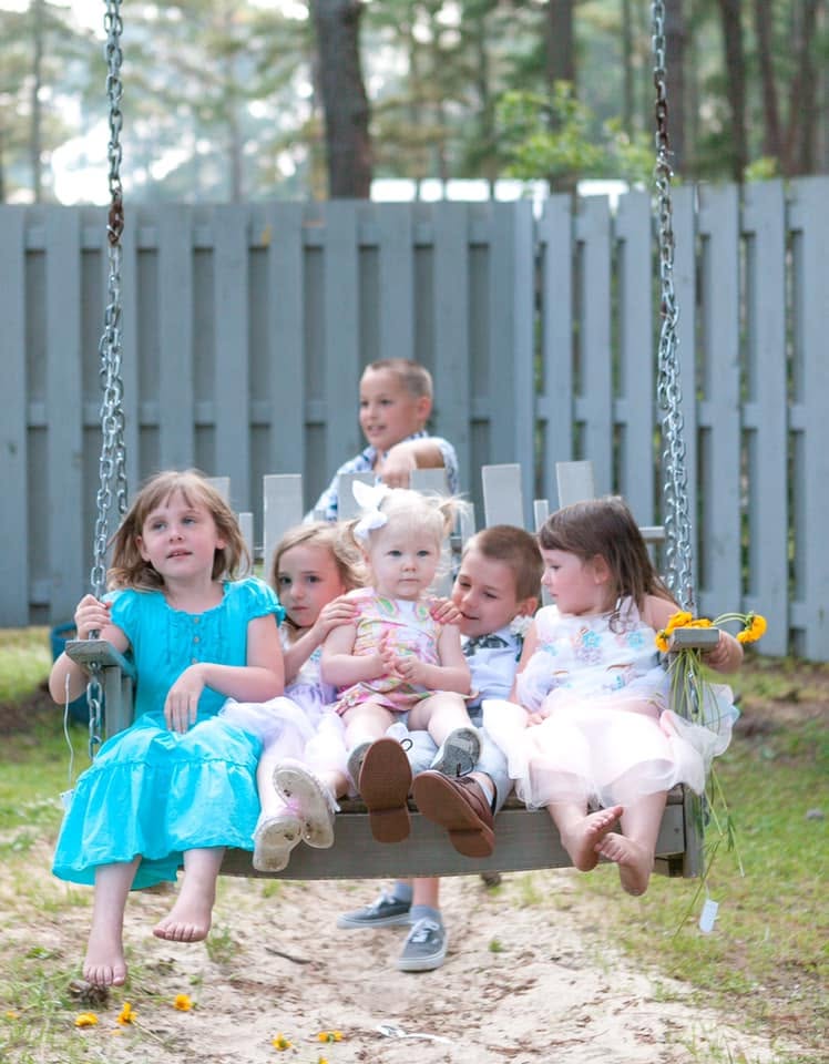 Six young children sitting together on the wooden porch swing at the Refuge Inn, under the pines.