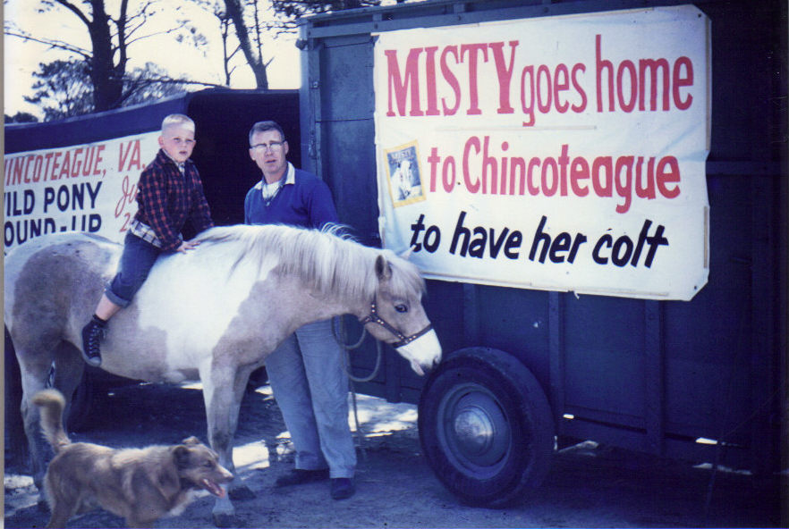 Carlton Leonard rides Misty of Chincoteague beside his father Don Leonard, in front of a trailer with a banner reading Misty goes home to Chincoteague to have her colt.
