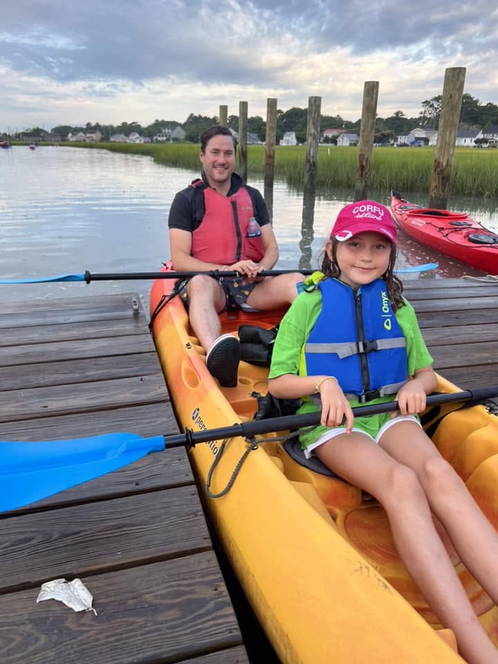 A man and a girl in life jackets sharing a kayak from Chincoteague Island Outfitters, beside the dock on Chincoteague Island.