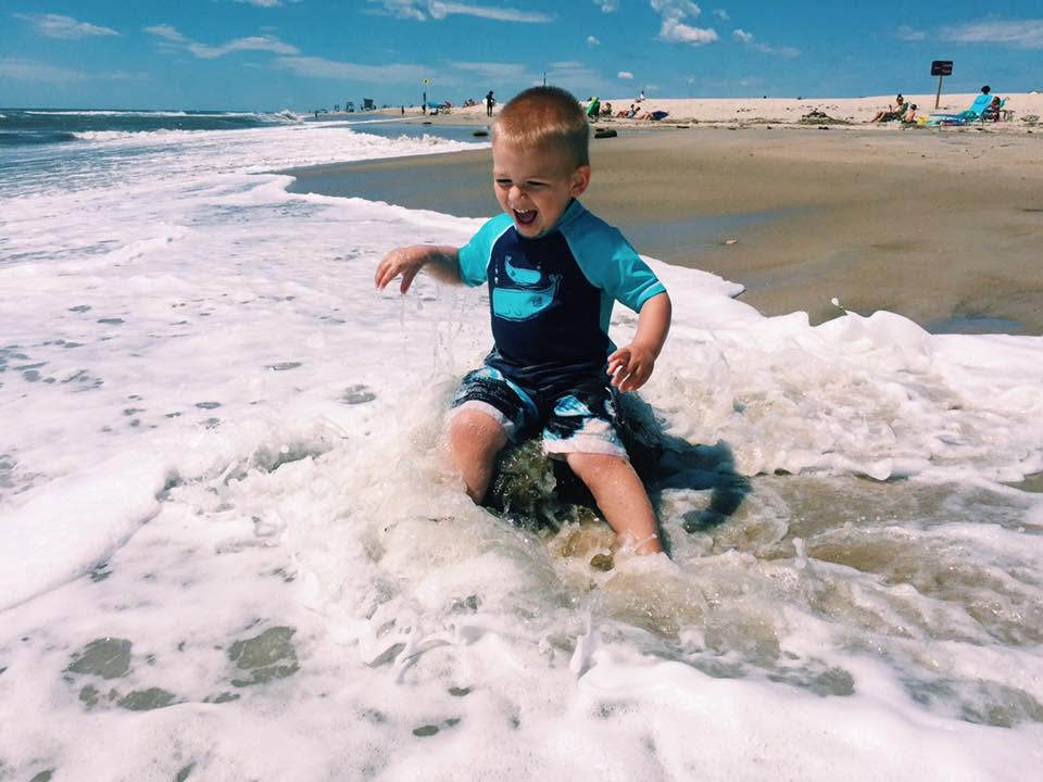 A toddler sitting in the shallow surf at the Assateague beach, laughing.