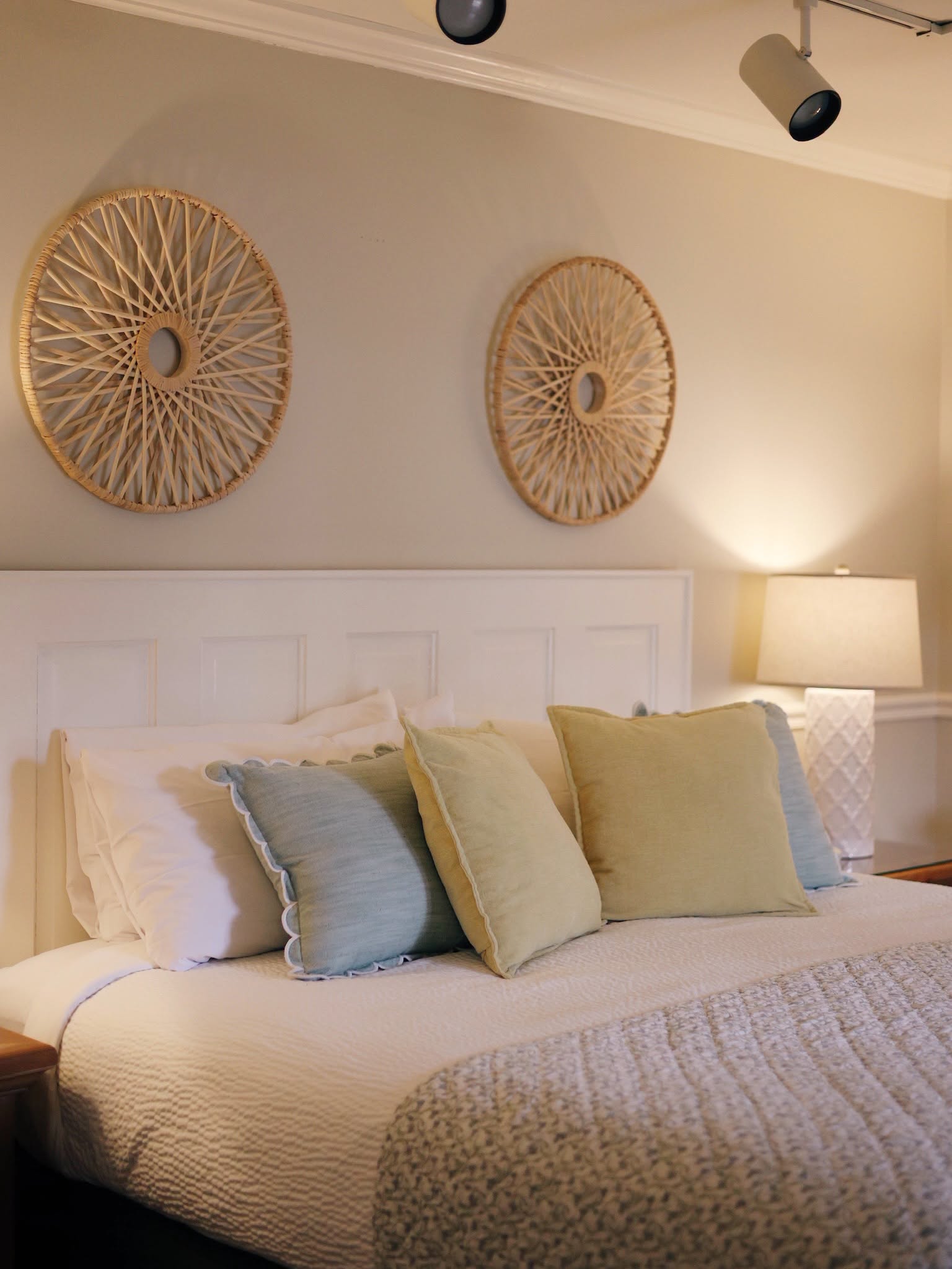 A made bed at the Refuge Inn with layered pillows and two woven rattan sunbursts on the wall above the headboard.