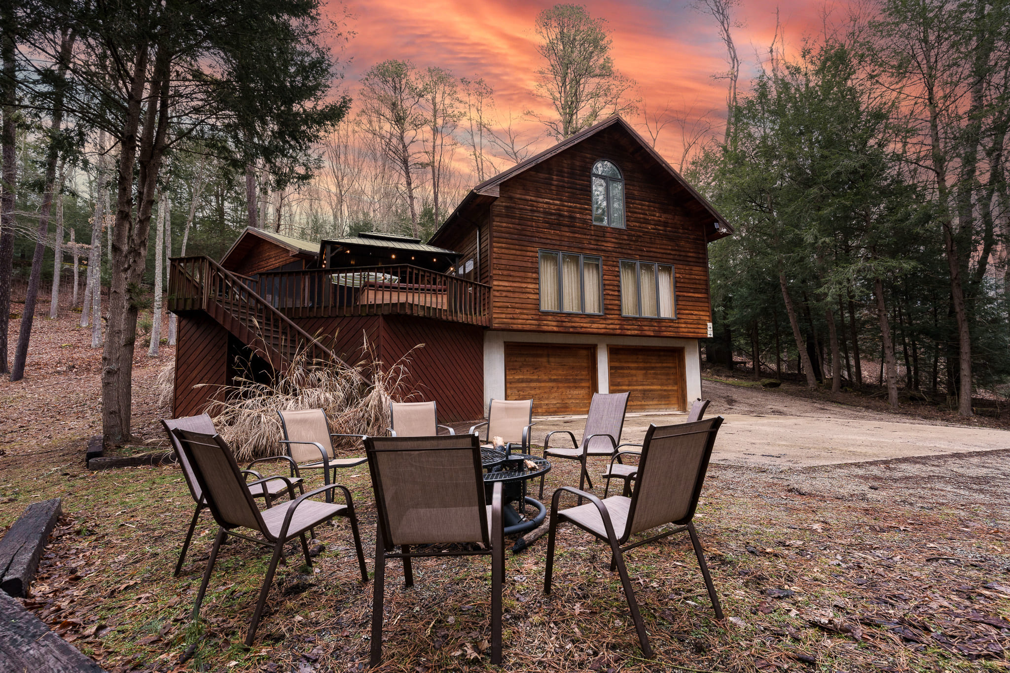 A wooden cabin surrounded by trees, featuring a seating area with chairs and a sunset sky.