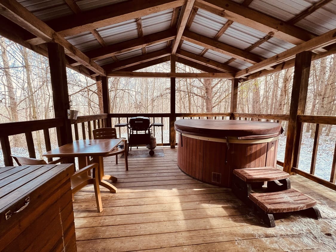 A wooden patio featuring a hot tub, a dining table, and a grill, surrounded by trees.