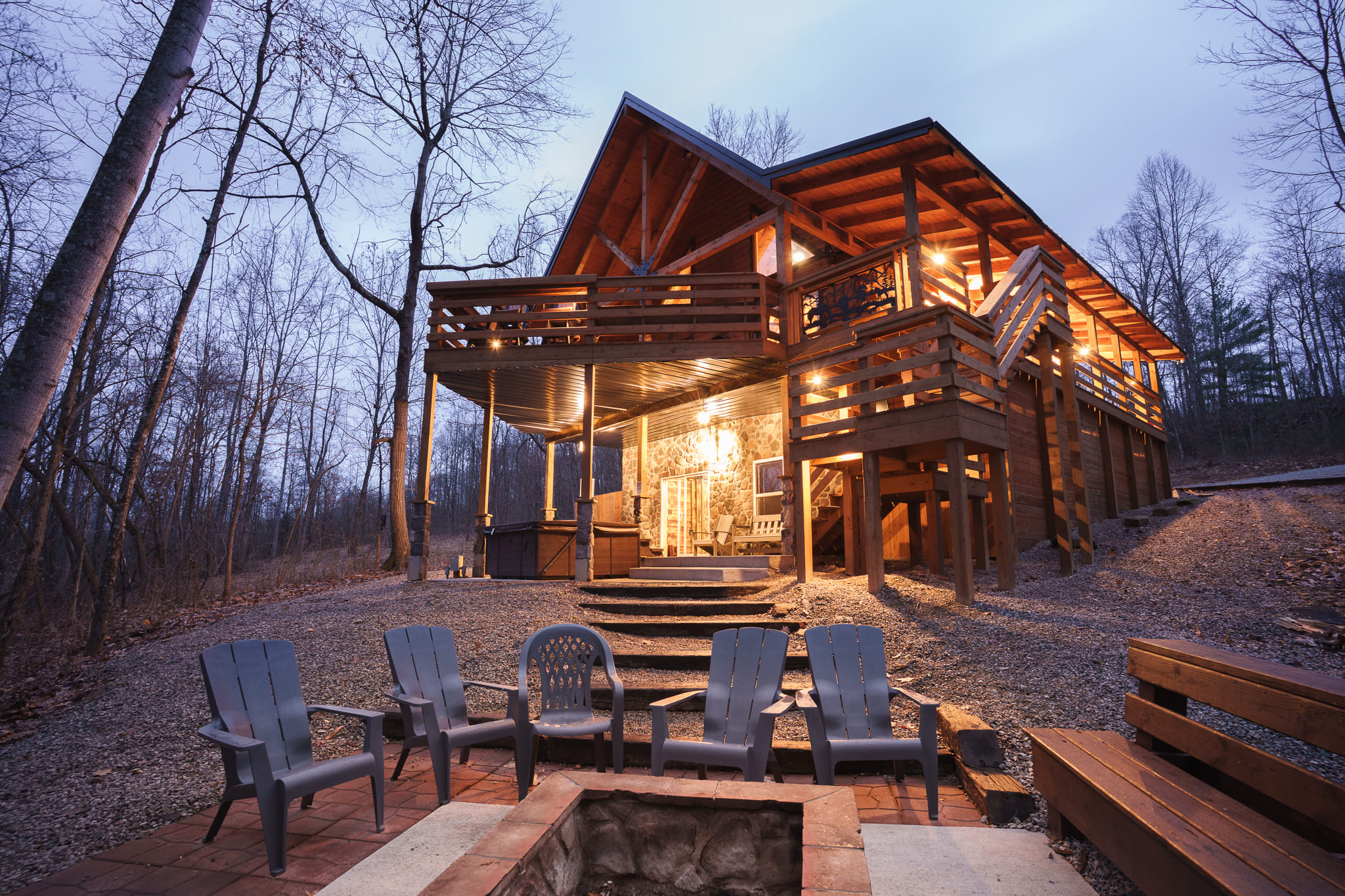 A sprawling wooden cabin with a spacious deck, surrounded by trees and featuring a fire pit with chairs in front.