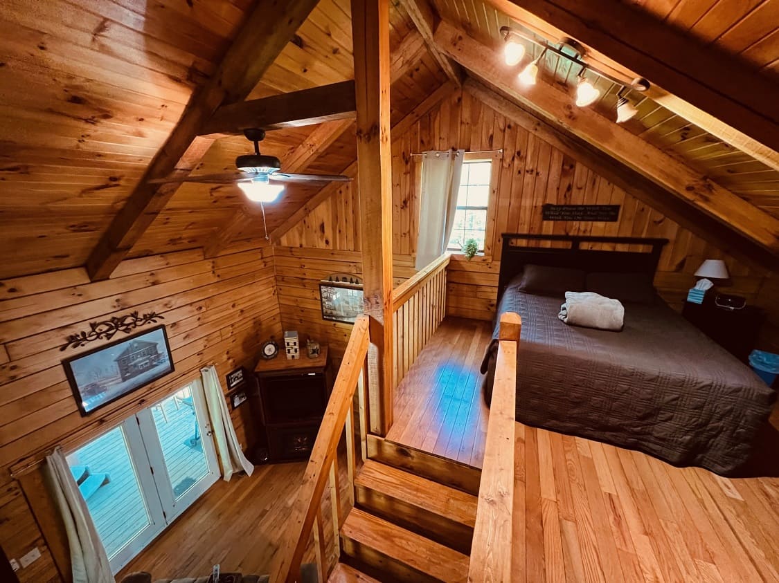A cozy loft bedroom with wooden walls, a queen bed, and a small staircase leading down to a living area.