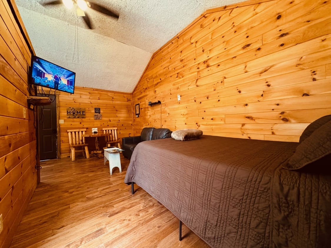 Cozy room with wooden walls, a bed, a small dining area, and a mounted TV.
