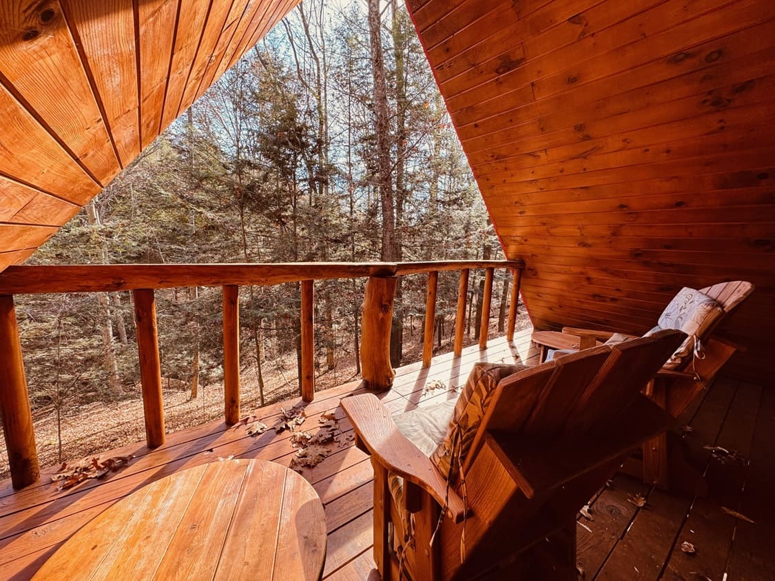 Wooden balcony overlooking a forest, featuring two chairs and a round table.