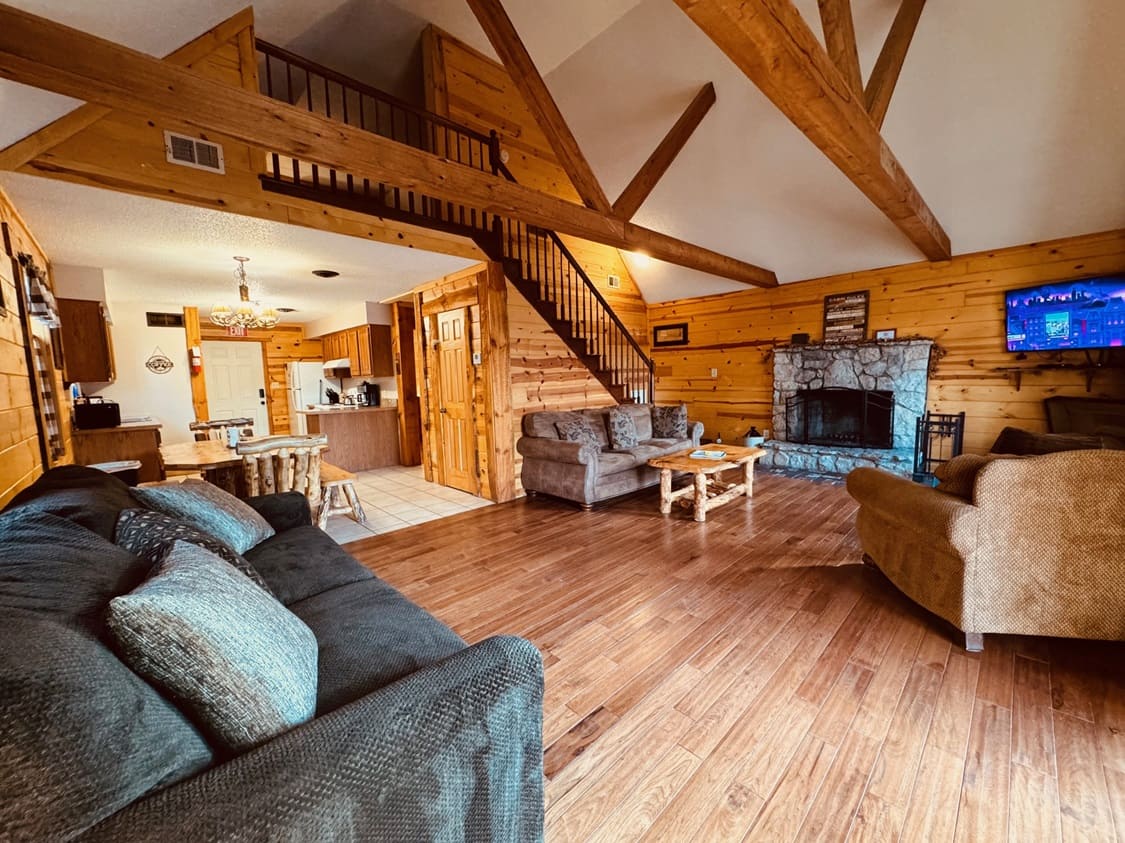 Open living area with wood-paneled walls, rustic furniture, a stone fireplace, and a staircase.