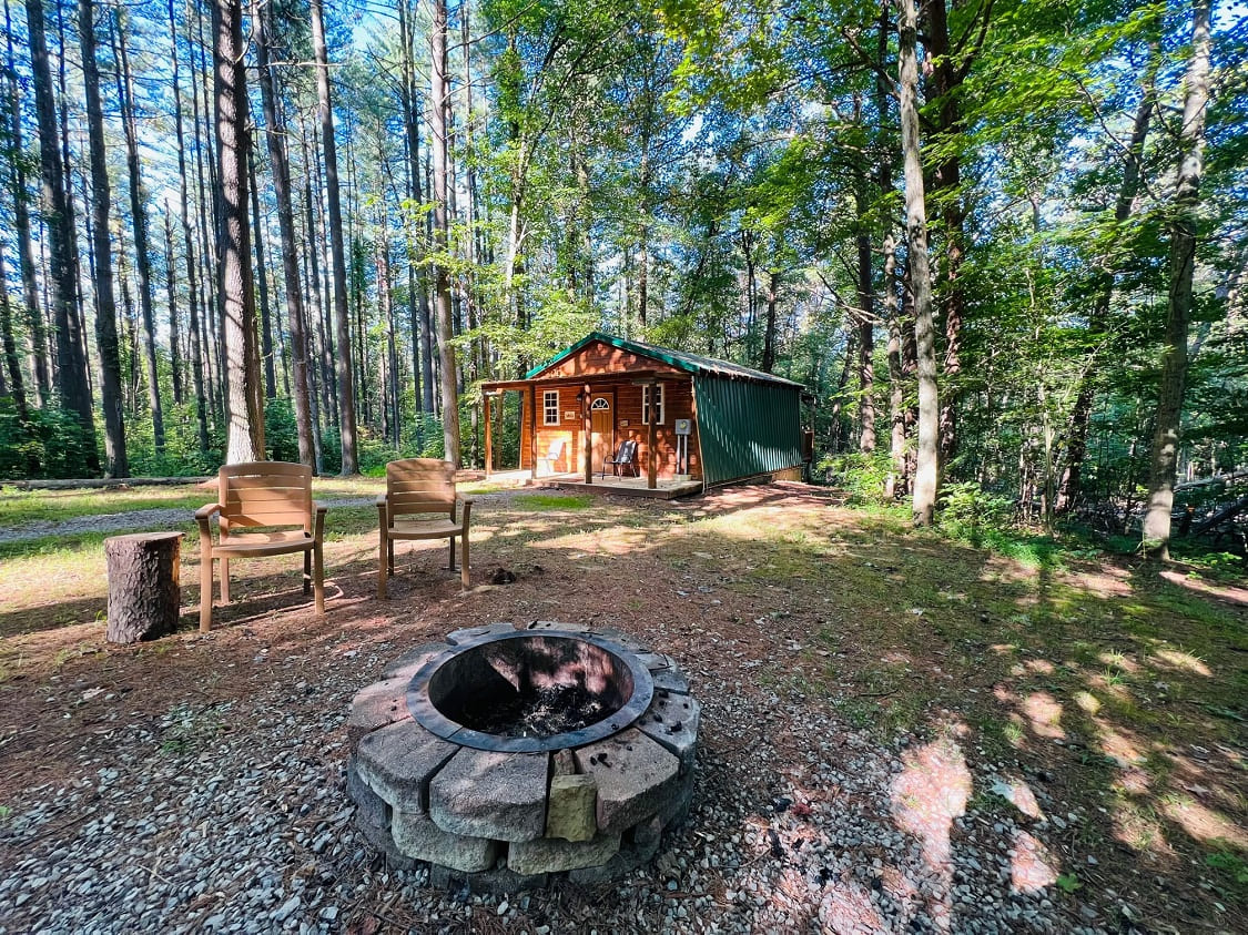 A cozy cabin surrounded by trees with two chairs and a fire pit in front.