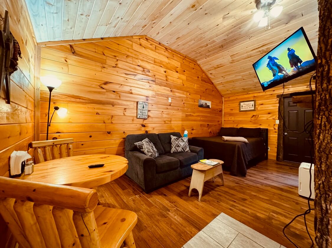 A wooden cabin perched on a hillside surrounded by bare trees under a colorful sunset sky.