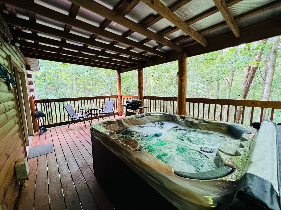 A hot tub on a wooden deck surrounded by trees, with a grill and seating area nearby.