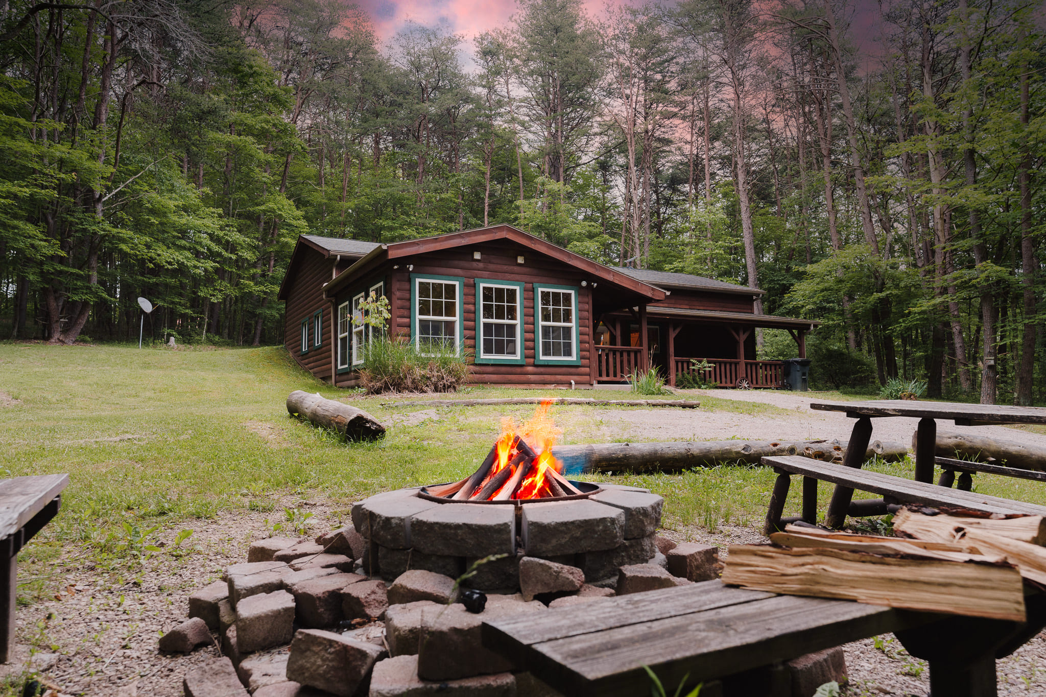 A log cabin surrounded by trees, with a fire pit in front.