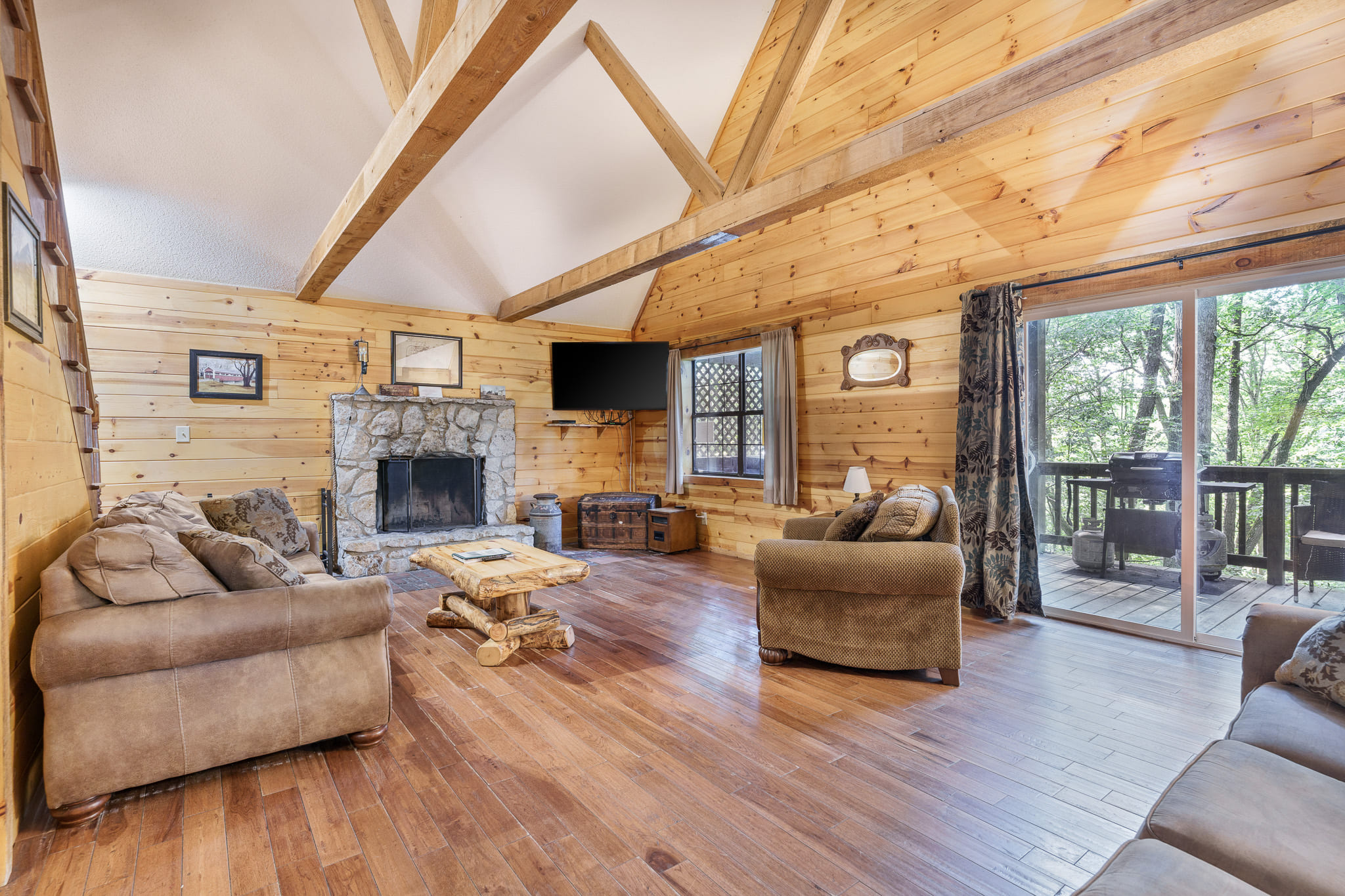 Cozy wooden cabin interior with a stone fireplace, comfortable seating, and large windows overlooking a deck and trees.