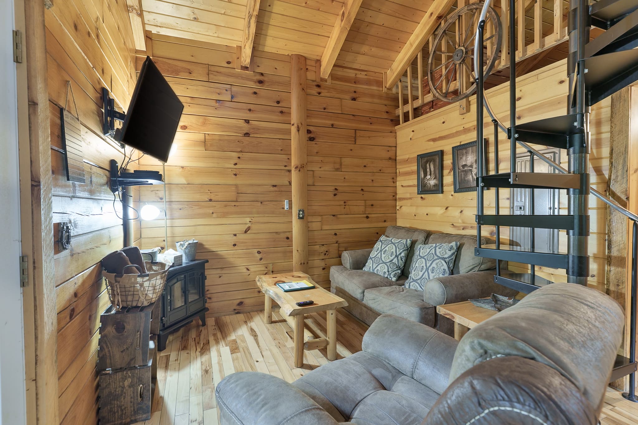 Cozy interior of a wooden cabin featuring a sofa, a small table, a television, and a spiral staircase.