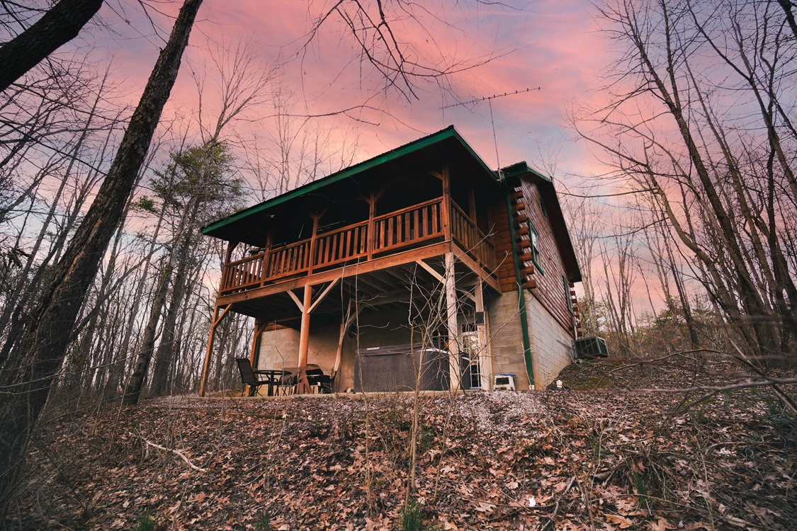 A wooden cabin on a hillside surrounded by bare trees under a colorful sunset sky.