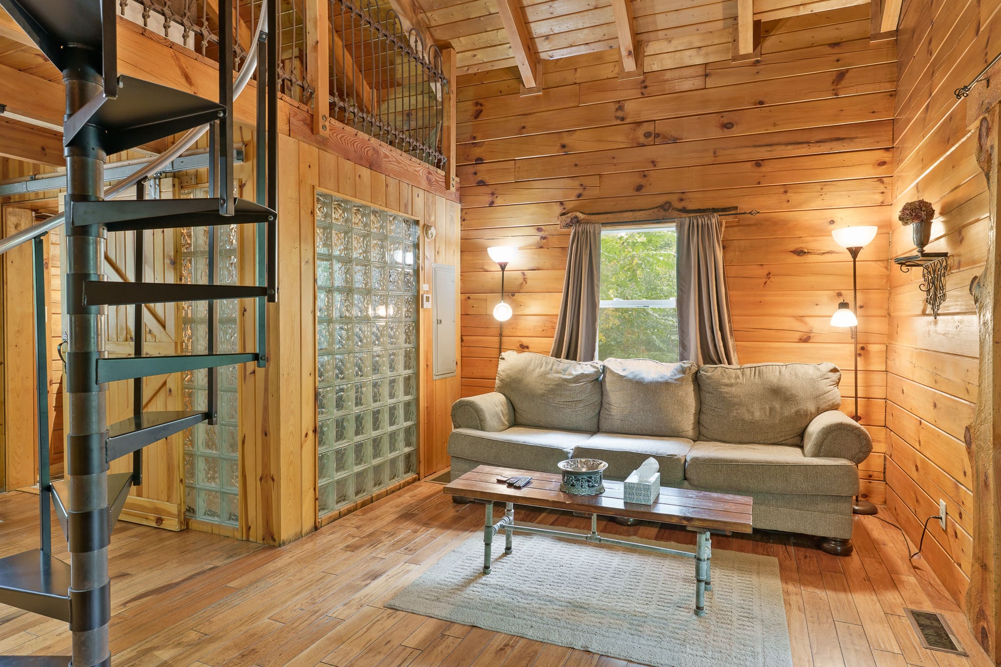 A cozy wooden living room features a spiral staircase, a comfortable couch, and natural light from windows.