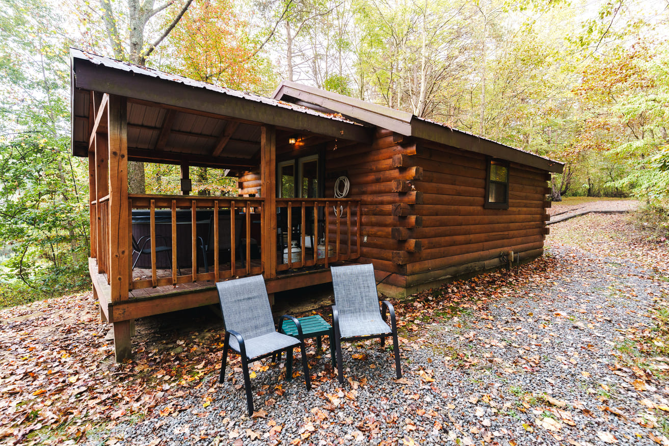 A rustic log cabin with a porch and two chairs sits amid autumn foliage.