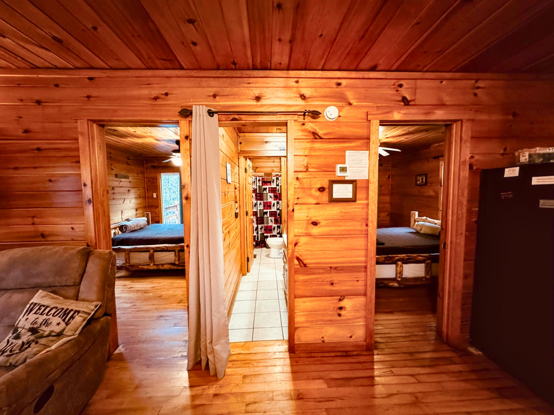 Interior of a wooden cabin featuring two doorways leading to bedrooms, a cozy couch, and a kitchenette.