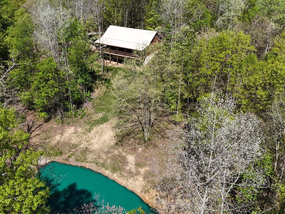 Aerial view of a cabin surrounded by green trees near a turquoise pond.