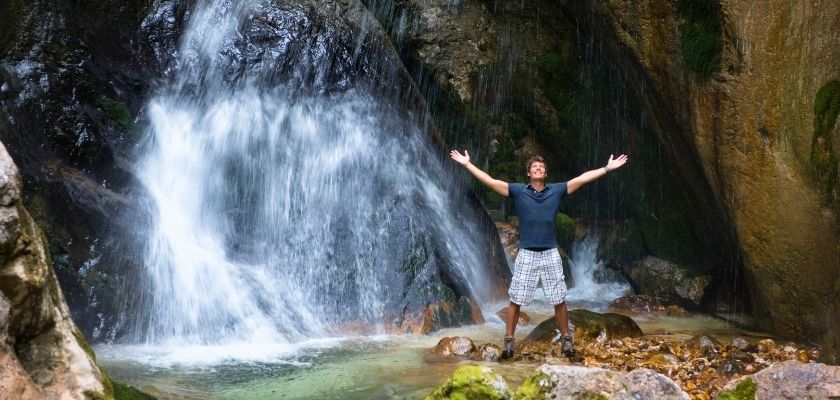 A waterfall cascades through a lush forest setting, surrounded by rocks and greenery, with a man standing in front with raised arms