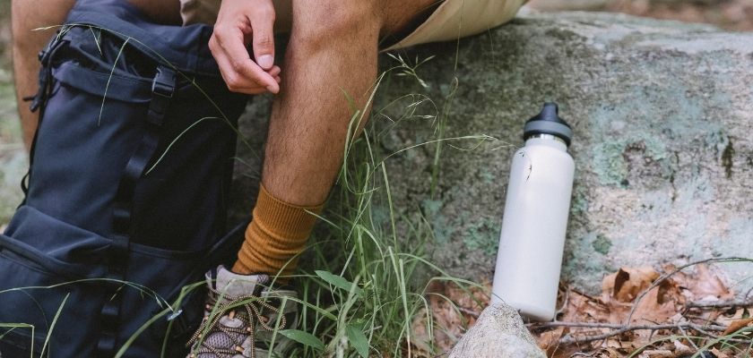 A close-up shows hiking gear beside a rocky trail, including a backpack, hiking boots, and a reusable water bottle.