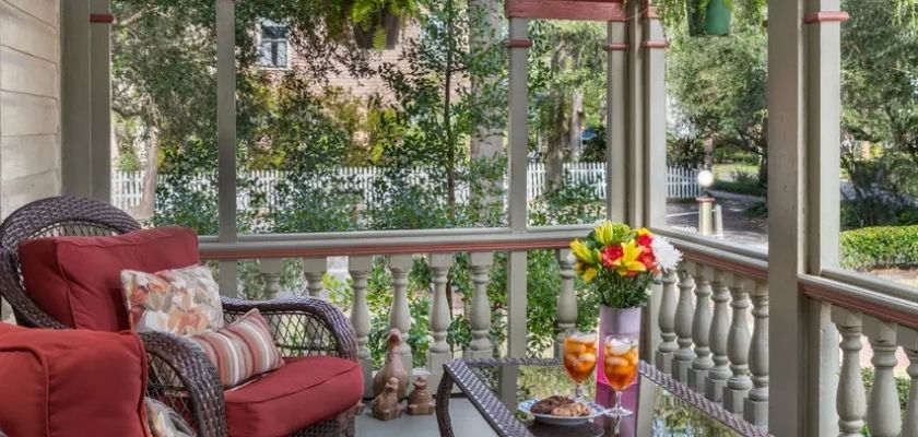Front porch of The Fairbanks House with cushioned chairs, a small table with drinks and flowers, and a view of a quiet tree-lined street.