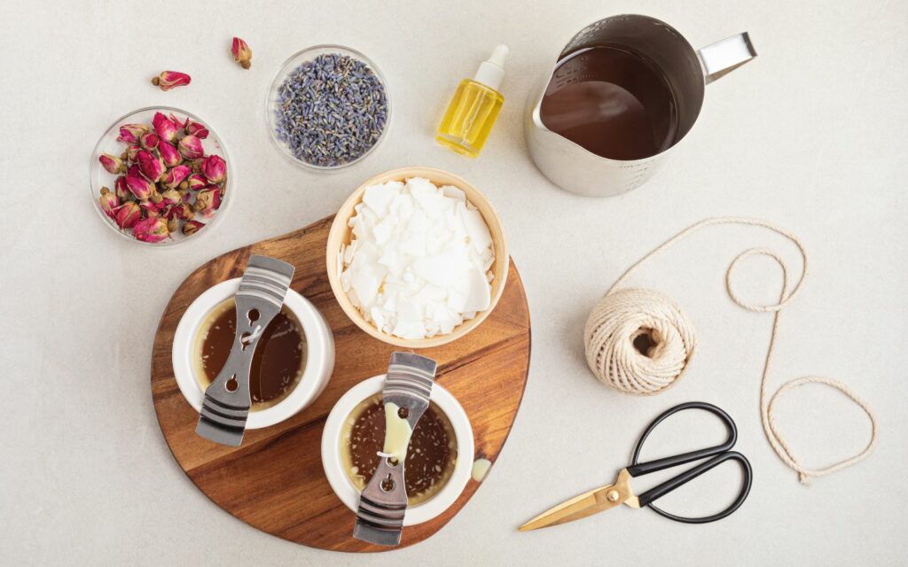 A wooden board with two candles in the process of cooling, wax ready to be melted, plus other candle-making supplies like wax string, scissors, essential oils, rose petals, and lavender buds.