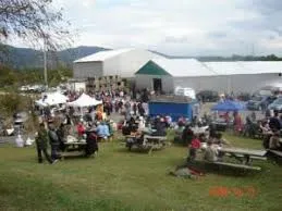 picnic tables set up in field with white barn in background and white tents with people walking around picnic tables set up in field with white barn in background and white tents with people walking around