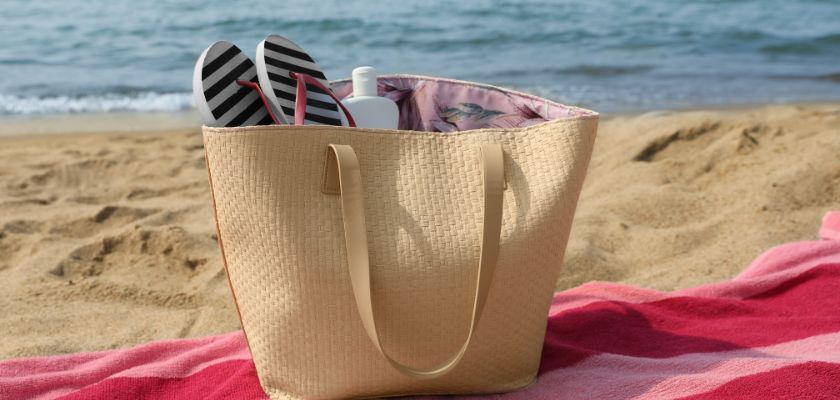 A beach bag with flip flops sitting on a beach blanket with Lake Michigan water lapping against the shore in the distance.