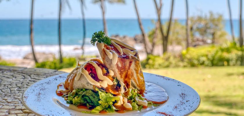 Plated wrap drizzled with sauce and garnished with greens set on a table overlooking a beach with ocean and palm trees in the background