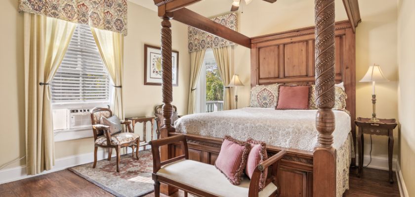 Room at the Kenwood Inn with a carved wooden four poster bed with patterned bedding, soft lighting, and classic furnishings near large windows