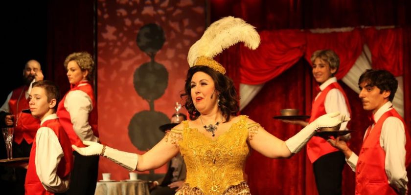 Performer in a gold costume and feathered headpiece singing on stage with servers in red vests holding trays in a theatrical setting